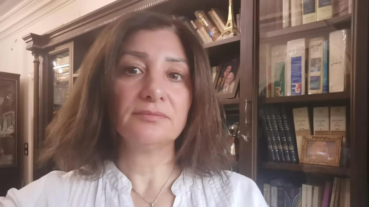 A woman with shoulder length brown hair and in a white shirt looks to camera with a book case behind her