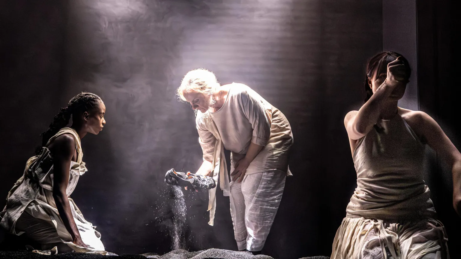 Raisa Šehu A scene in a play with three actors dressed in minimalist, loose clothing. A woman in the left is crouched watching another woman pulling a sandal from a piles of black dust. To her right another woman is holding an outstretched fist to the camera.